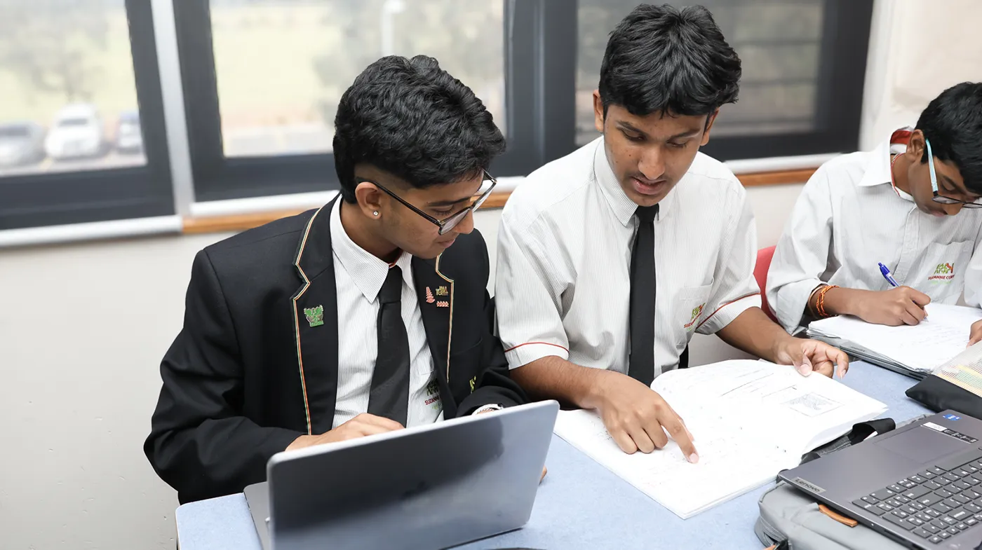 Two students in school uniforms sit at a desk, one pointing at a book. A laptop is nearby. Another student writes notes in the background.