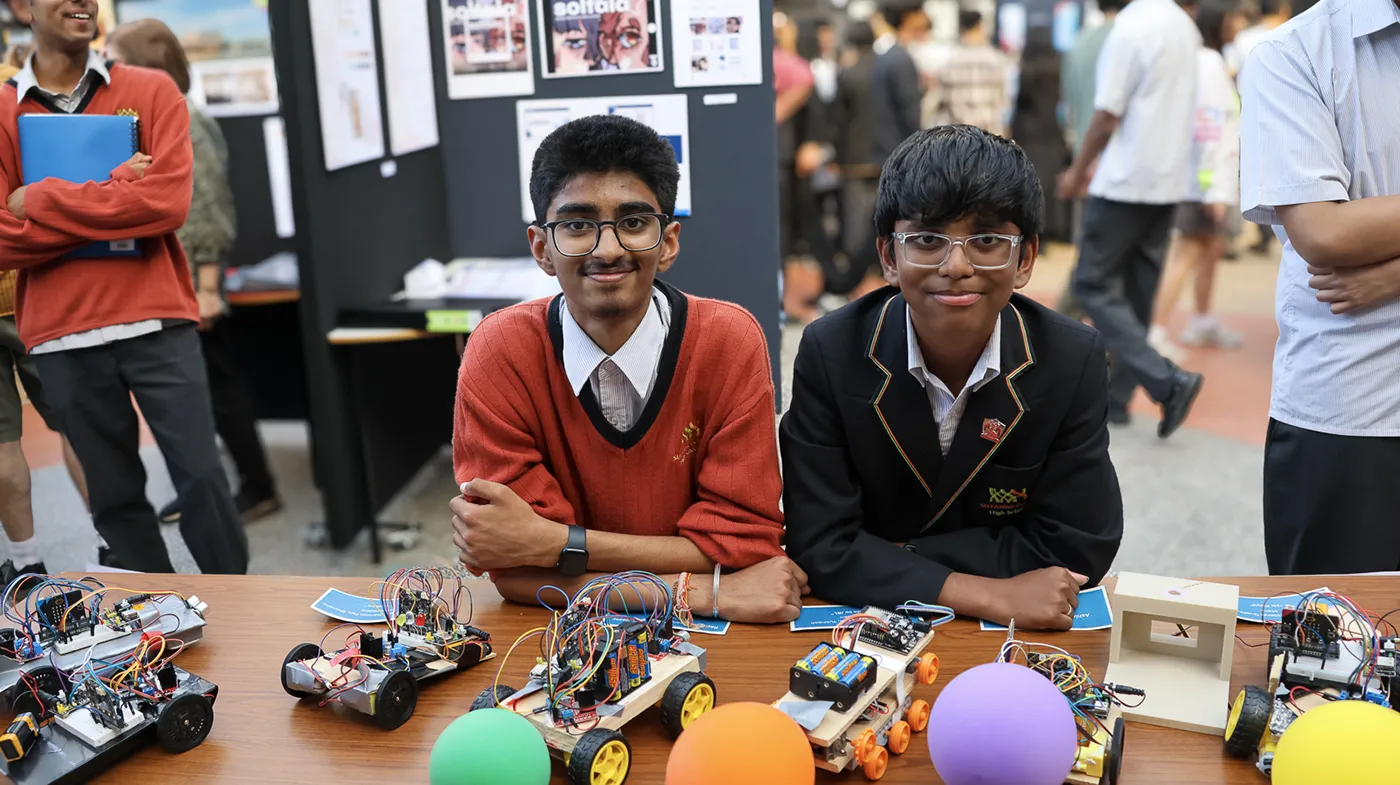Two students sit behind a table displaying robotic projects with electronic components. They are wearing school uniforms, and the background shows other people and posters.