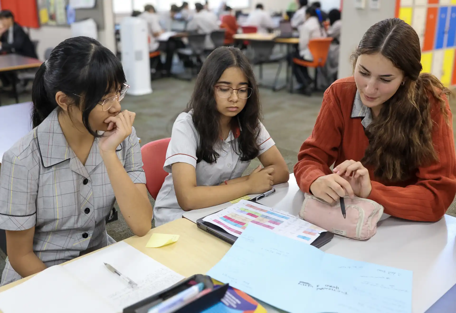 Three students sit around a table in a classroom, discussing work. They have notebooks and papers with colorful charts. One student gestures with a pen. The focus is on collaboration and learning.