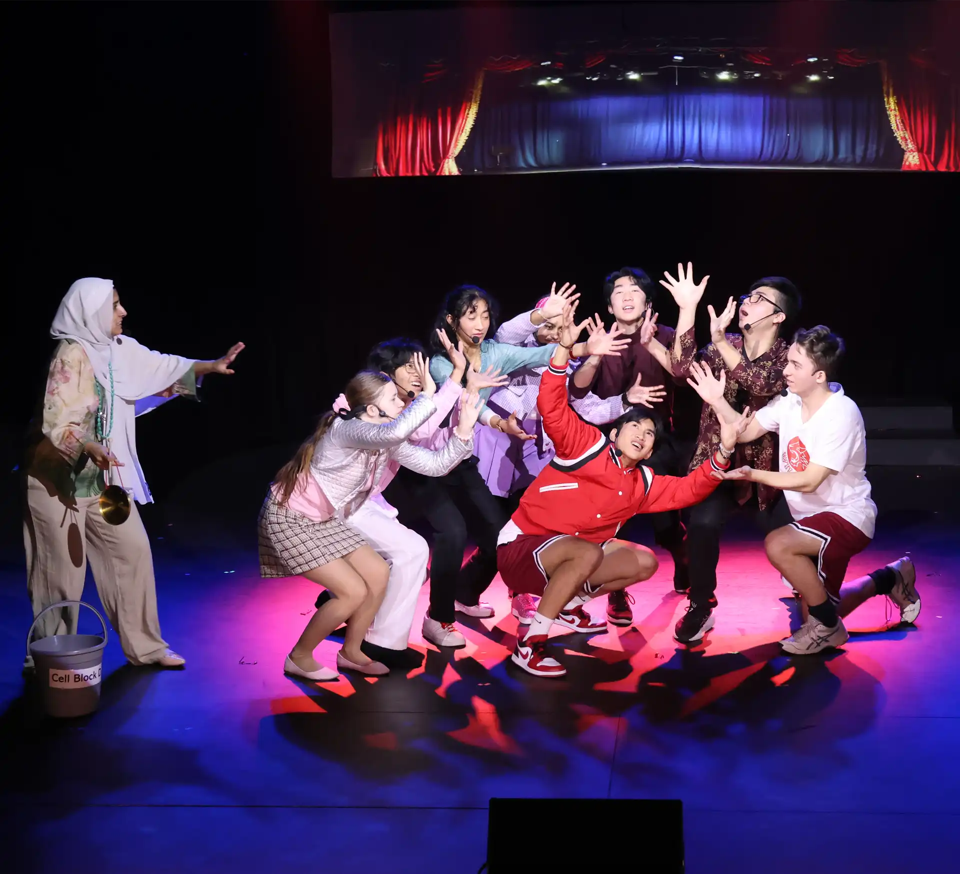 A group of performers on stage expressively reaching towards a spotlight. They are dressed in colorful, casual clothes. A person on the left, wearing a headscarf, holds a bucket and looks towards the group. A dramatic backdrop features a stage curtain.