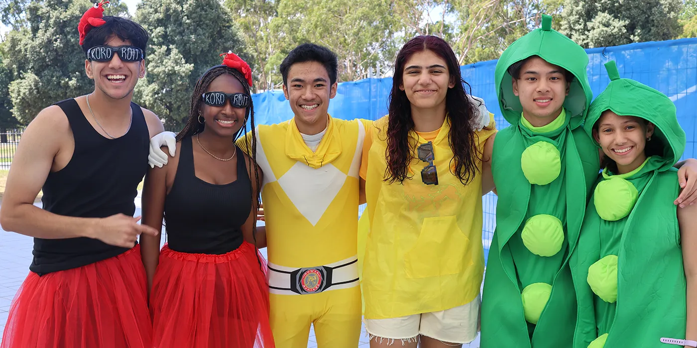 A group of six students in colourful costumes, including sunglasses, a red tutu, a yellow superhero suit, and pea pod outfits, stand smiling together outdoors.