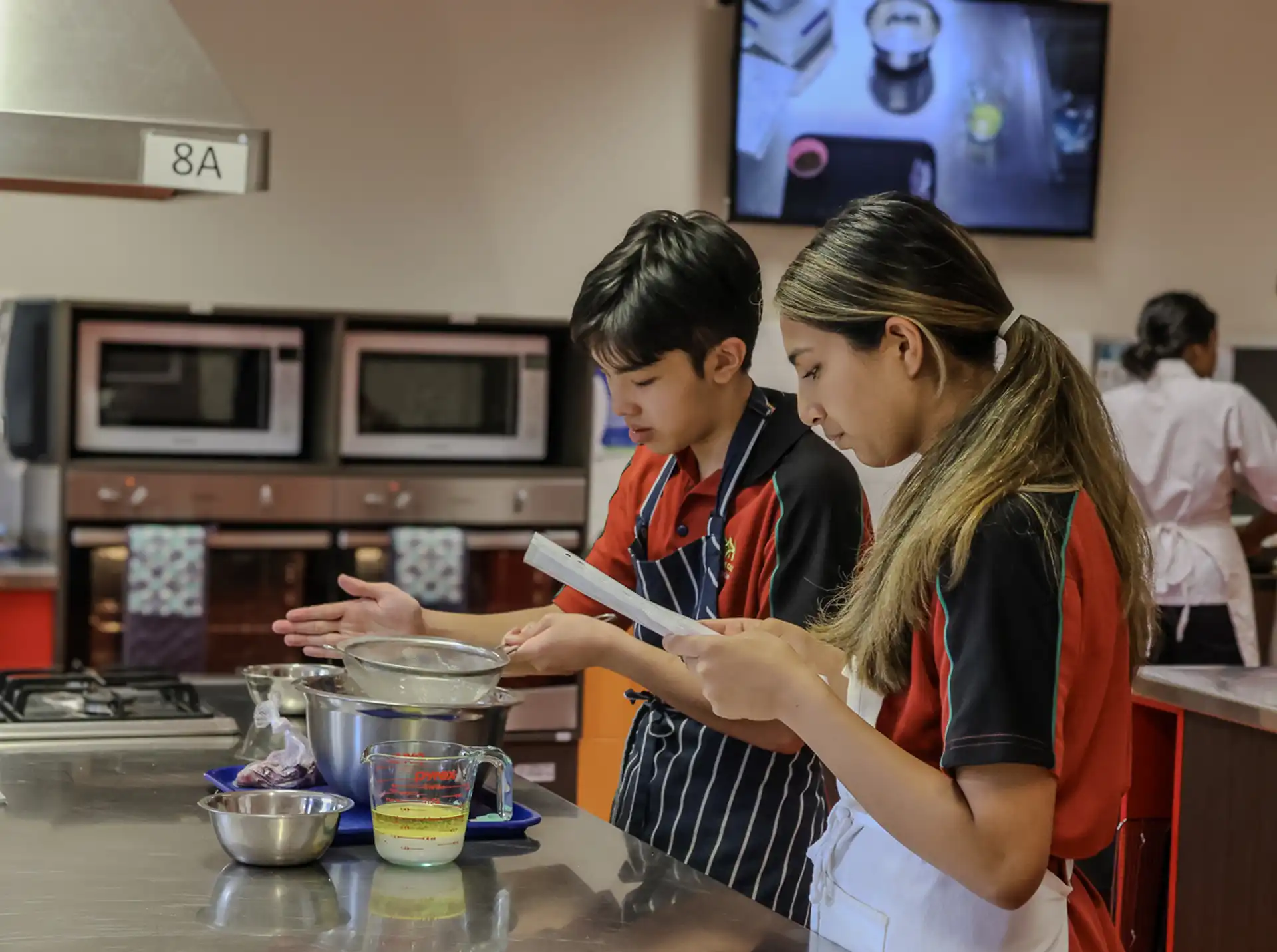 Two students in a kitchen classroom focus on a cooking task. The boy stirs ingredients in a pot, while the girl reads from a paper. They wear red and black uniforms with white aprons. Microwaves are visible in the background.