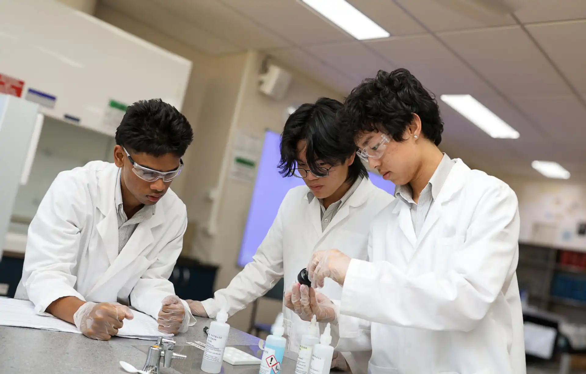 Three individuals in lab coats and safety goggles conduct an experiment in a laboratory setting. They are focused on mixing chemicals in bottles, with additional lab equipment in the background.