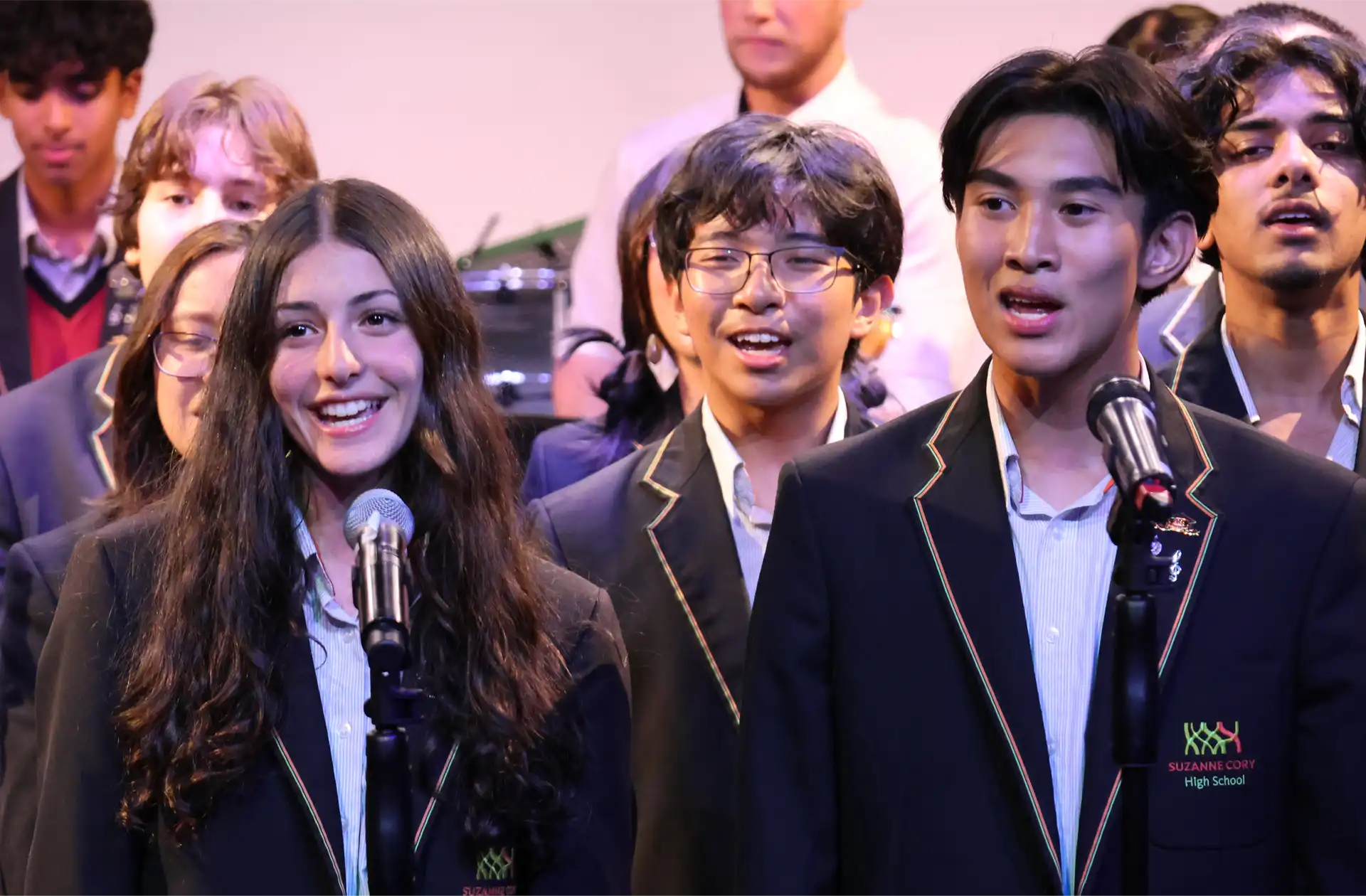 A group of students in school uniforms is singing on stage. Two students stand in front, smiling and singing into microphones. They wear blazers with embroidered school emblems. A blurred band is visible in the background.