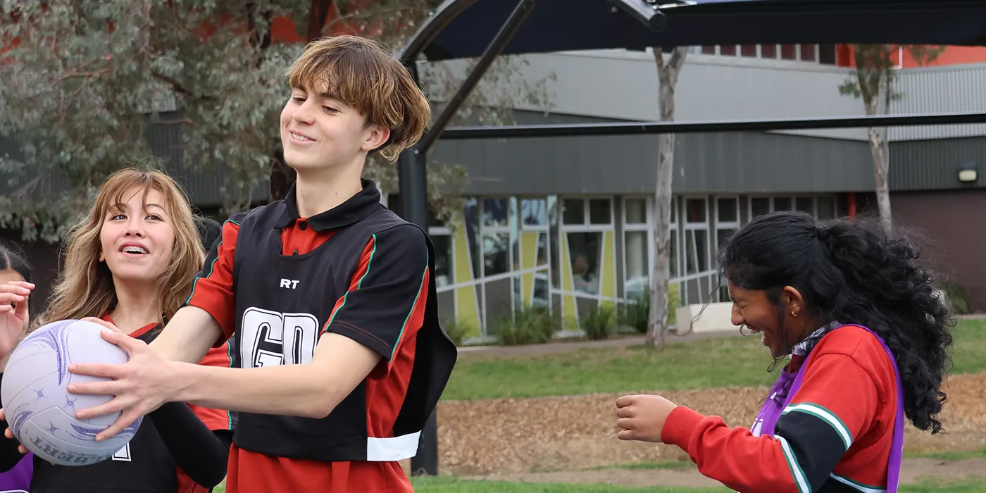 Three people outdoors playing netball, with one holding the ball. They're wearing sports attire and appear engaged and happy, with trees and a building in the background.
