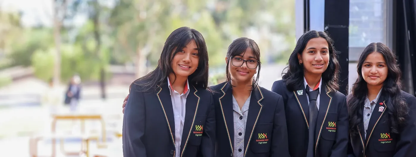 Four students in matching school uniforms pose for a photo outdoors, smiling. They wear black blazers with school badges on the chest. Blurred background of trees and an outdoor setting is visible.